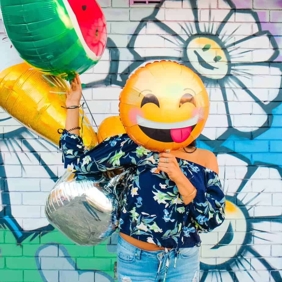 A person in a flowered shirt holding a smiley face balloon over her face, standing in front of a flower graffiti wall.